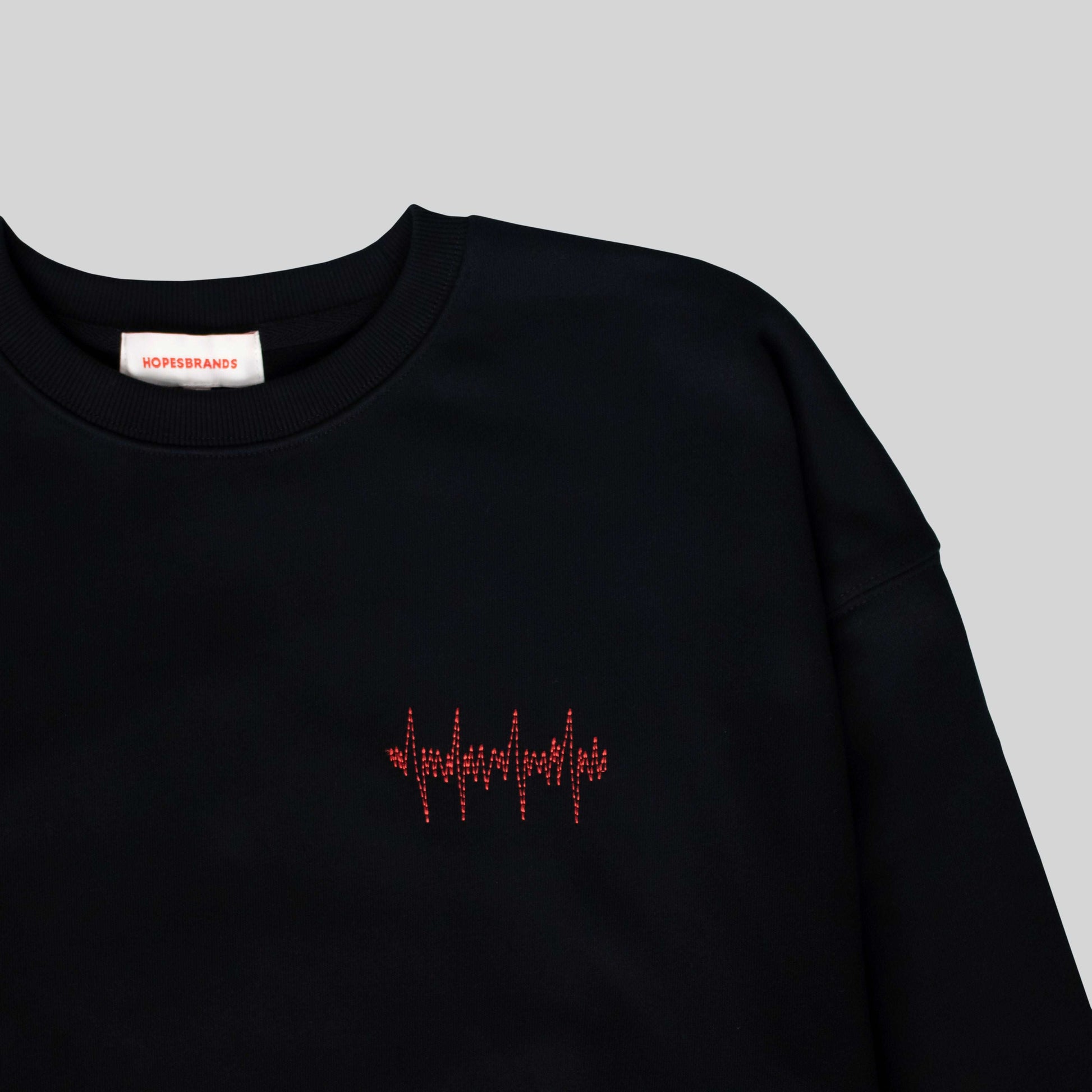 Close-up of the Hopesbrands Big Heart crewneck sweatshirt in black, showing the red lifeline stitch logo on the left chest. Made from a comfortable cotton blend.