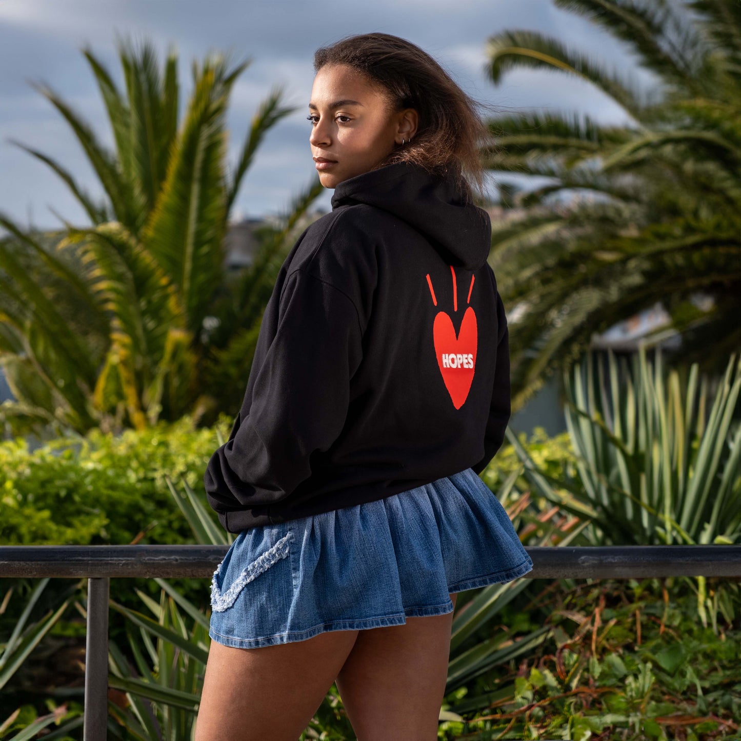 Female model wearing black Big Heart hooded sweatshirt from Hopesbrands, set against a beach backdrop.