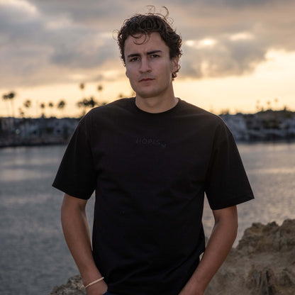 Male model wearing Hopesbrands black men's tonal short sleeve T-shirt with tone-on-tone 'Hopes' logo, set against a sunset beach backdrop. Emphasizes casual style and unisex appeal.