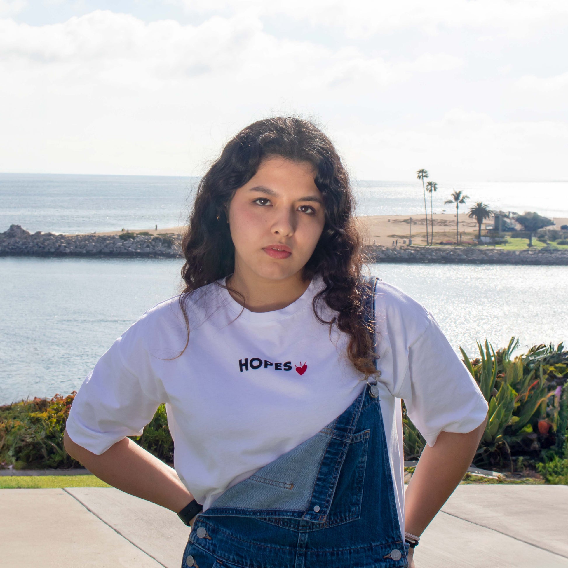 Model wearing a white Hopesbrands Cool Touch T-Shirt with embroidered 'Hopes' logo, standing by the sea. The relaxed fit showcases durability and timeless design, ideal for everyday wear.