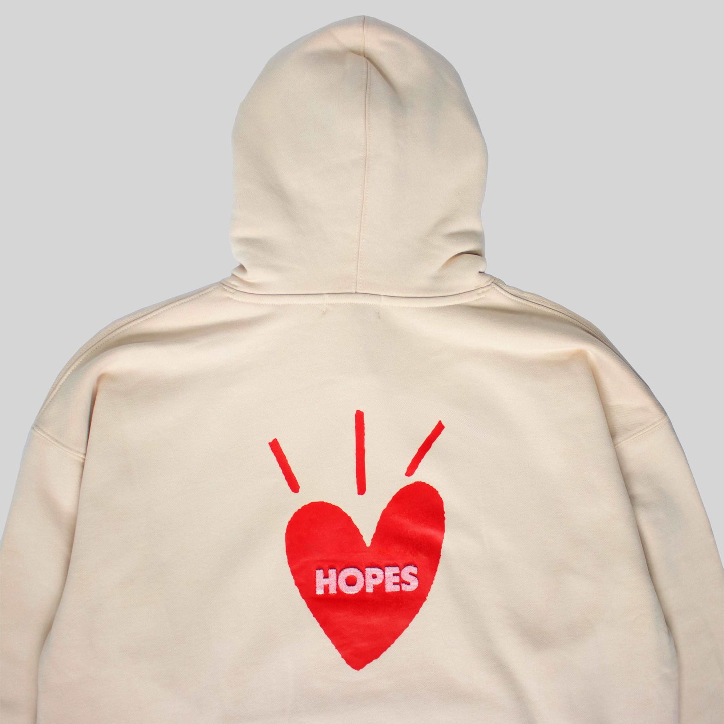 Close-up of tan Big Heart hooded sweatshirt with printed Big Heart logo and red-toned toothbrush embroidery on 'Hopes' letters.